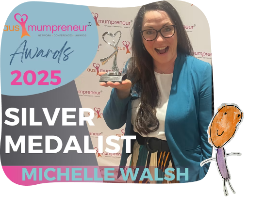 Woman in a blue blazer smiles and holds a silver trophy, standing in front of a banner reading “2025 Ausmumpreneur Awards Silver Medallist Michelle Walsh.”.