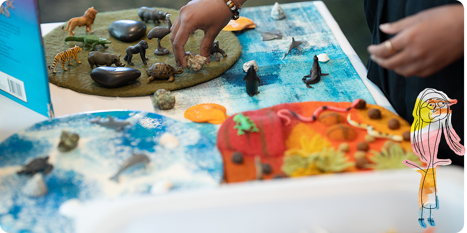 A person arranges animal figurines on felt mats representing land and water habitats; a colourful cartoon character is illustrated in the lower right corner.