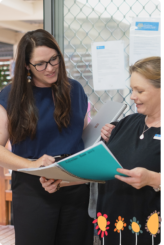 Two women stand indoors, looking at documents together. One holds a laptop, and the other holds a large blue folder. Papers are posted on a glass door in the background.