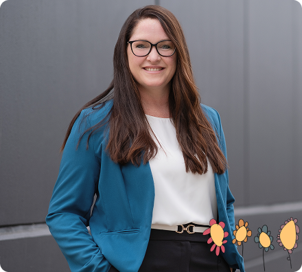 Woman with long brown hair, glasses, and a blue blazer stands in front of a grey wall, smiling. Colourful flower graphics appear in the lower right corner.