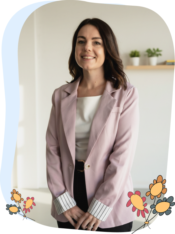 Woman in a light pink blazer stands indoors, smiling at the camera. She has shoulder-length brown hair. Cartoon flowers decorate the image corners. Shelf with small plants is in the background.