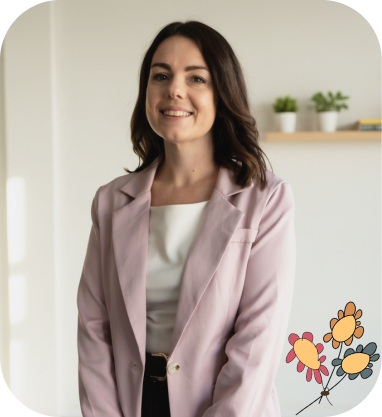 Woman with shoulder-length brown hair wears a light purple jacket and white top, standing indoors with plants on a shelf in the background. A floral graphic is in the lower right corner.