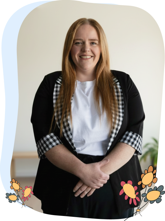 Woman with long red hair wearing a black blazer with checked cuffs and a white shirt, standing and smiling. Simple background with illustrated flowers at the bottom of the image.