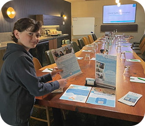 A person stands beside a conference table set with papers, glasses, and pens in a meeting room, holding a brochure and looking towards the camera.