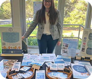 A woman stands behind a table displaying brochures, leaflets, and informational materials at an indoor event with large windows in the background.