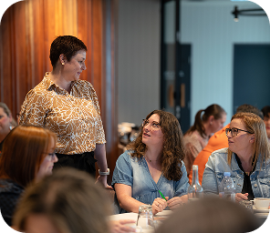 A woman stands and talks to two seated women at a table during a meeting or workshop, with other people visible in the background.