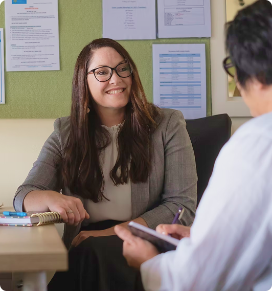 Two people sit opposite each other in an office, one smiling and listening whilst the other takes notes in a notebook. Documents are pinned on the wall behind them.