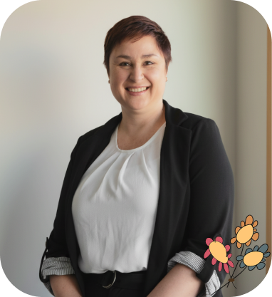 Smiling person with short brown hair wearing a white blouse and black blazer, standing against a light background with a small illustrated flower graphic in the bottom corner.