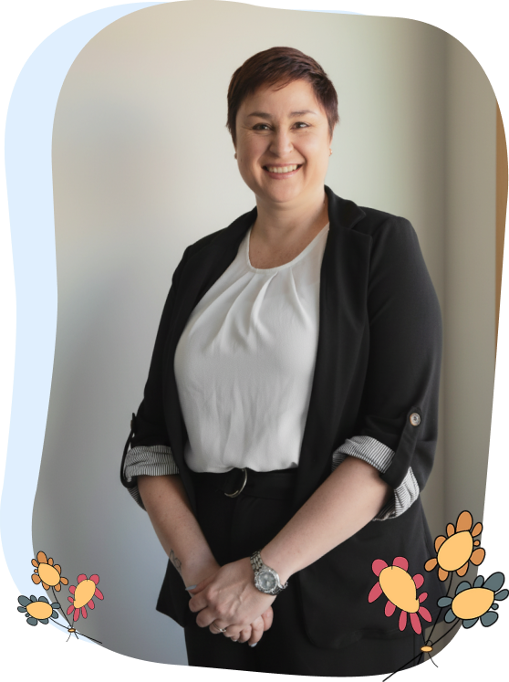 A person with short brown hair, wearing a black blazer and white blouse, stands indoors smiling. Decorative floral graphics are at the corners of the image.