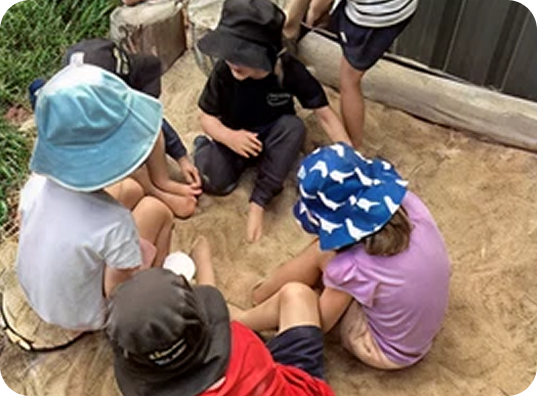 Five children wearing hats sit and play together in a sandy area outdoors near a building.