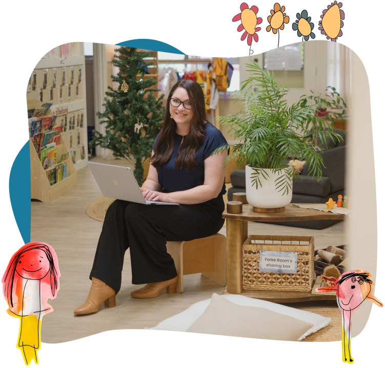 A woman sits on a small wooden chair using a laptop in a classroom decorated with plants, children's artwork, and a Christmas tree.
