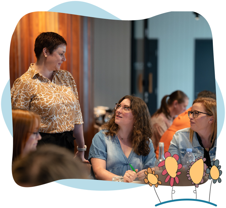 A woman standing speaks to two seated women at a table during a group meeting. The setting appears informal and collaborative, with people engaging in conversation. Flowers decorate the image border.
