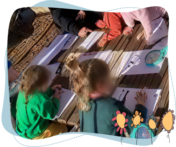 Children sit around a wooden table outdoors, drawing on white paper with pencils. Magnifying glasses are on the table. Faces are blurred for privacy.