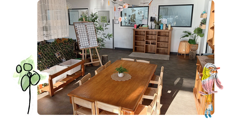 A classroom with wooden tables and chairs, shelves with storage boxes, a whiteboard, potted plants, and large windows letting in sunlight.