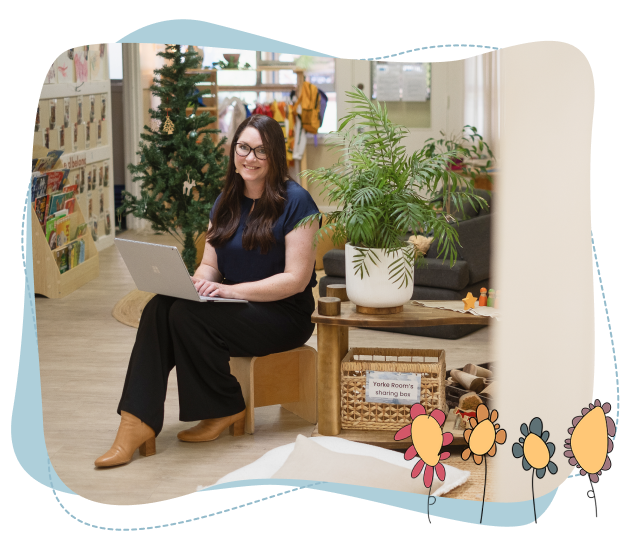 A woman sits on a small wooden chair using a laptop in a classroom decorated with a Christmas tree, books, and potted plants.