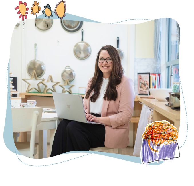 Woman with long brown hair and glasses sits at a table with a laptop in a kitchen, smiling. Pots, pans, and star decor are visible on the wall behind her. Colourful drawings frame the image.