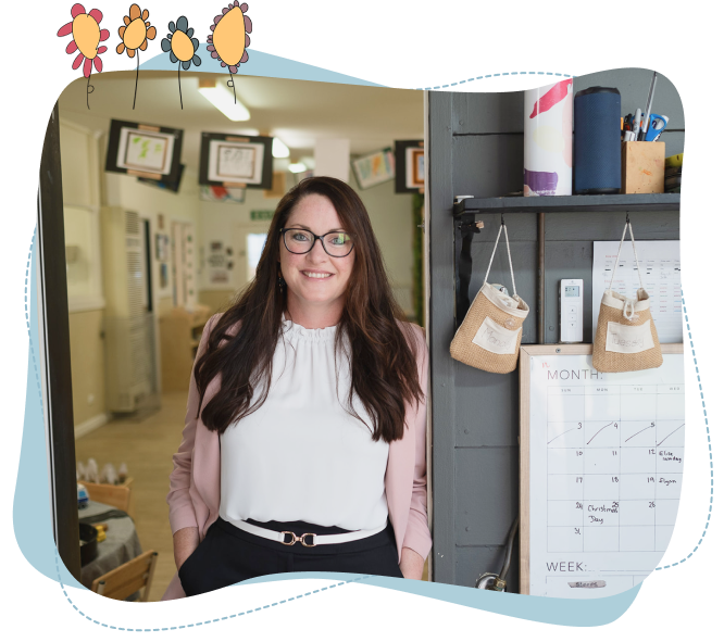 A woman with long brown hair and glasses stands smiling in an office doorway, next to a wall calendar and hanging pouches.