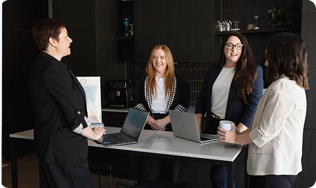 Four women in business attire stand around a table with laptops, talking and smiling in a modern office setting.