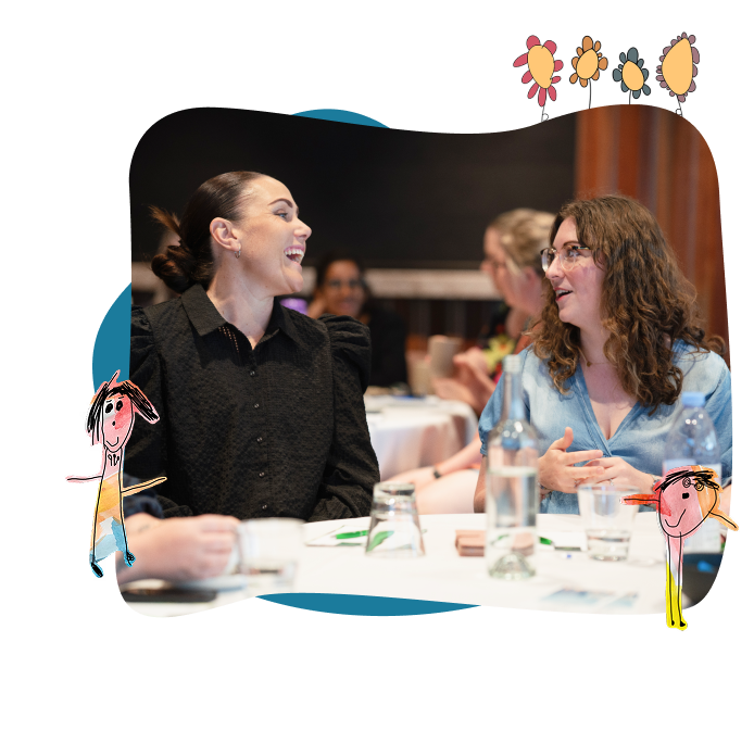 Two women sit at a table in conversation during an indoor event, with drinks and papers on the table and cartoon drawings surrounding the image.