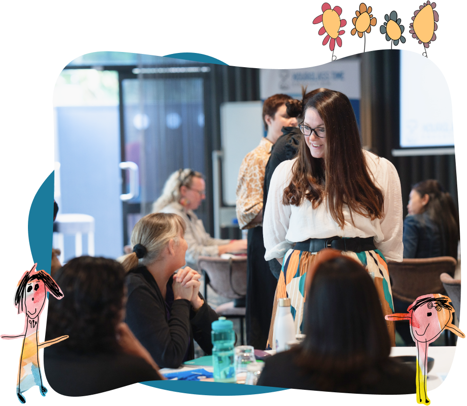 A woman stands and smiles whilst talking to seated colleagues in a meeting room, with childlike drawings and flowers framing the image.