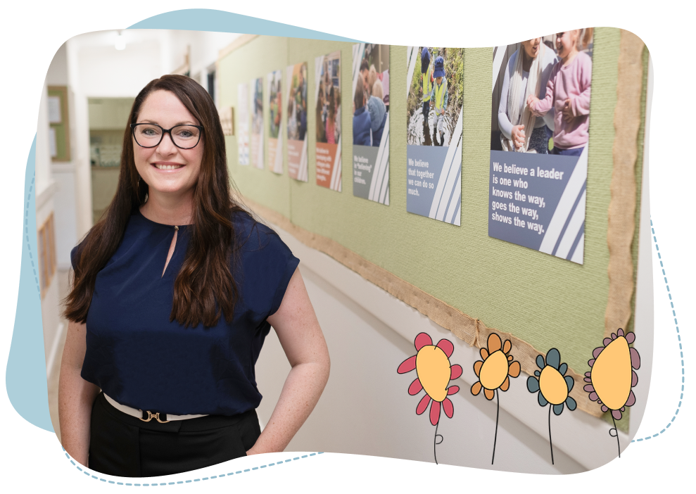 A woman with long brown hair and glasses stands in a corridor with motivational posters on a noticeboard behind her.