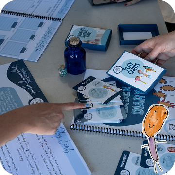 People interact with educational materials on a table, including playing cards, booklets, a blue bottle, and illustrated sheets with text and drawings.
