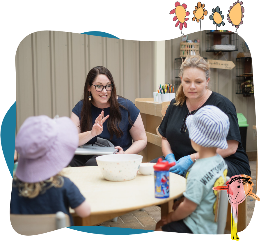 Two adults sit at a table with two young children wearing hats. One adult gestures with her hand while the other looks on. Art supplies and shelves are visible in the background.