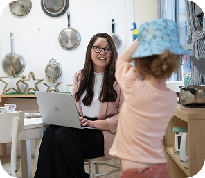 A woman working on a laptop at a kitchen table smiles at a child wearing a blue hat who is standing nearby.