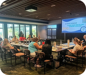 People seated around tables in a conference room attending a presentation, with a large screen displaying meeting information at the front of the room.