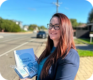 Woman with long brown hair and glasses stands on a pavement, holding a closed laptop and ID badges, with a street and houses visible in the background.