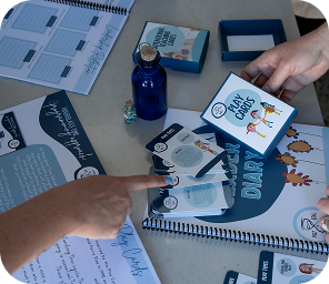 Two people interact with a set of “Play Cards”, a blue glass bottle, dice, and a spiral-bound “PLAY DIARY” on a table, suggesting a tabletop activity or game in progress.